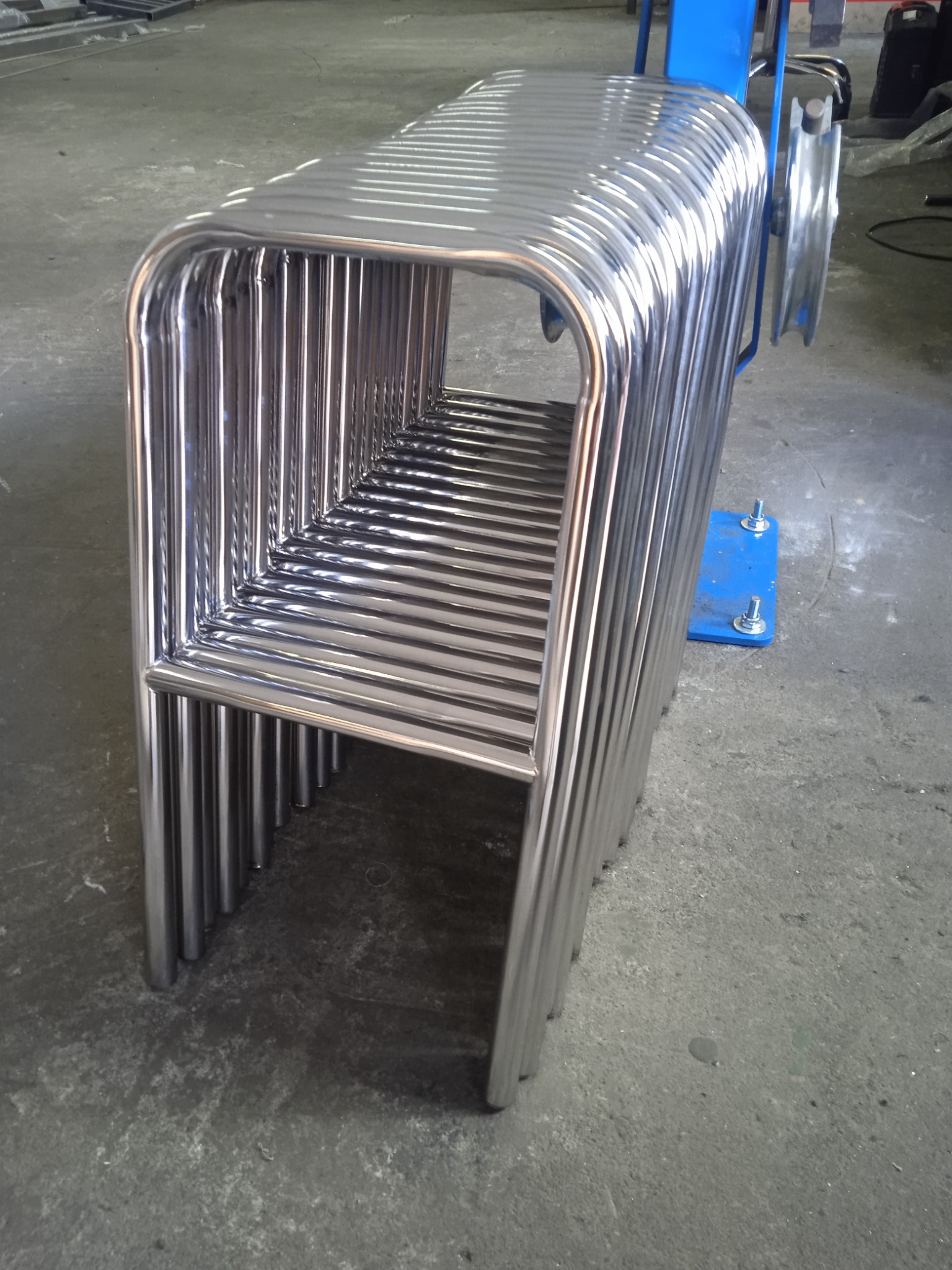 Stos wygiętych, wypolerowanych rur ze stali nierdzewnej, ułożonych jedna na drugiej, obok niebieskiego urządzenia do gięcia rur.