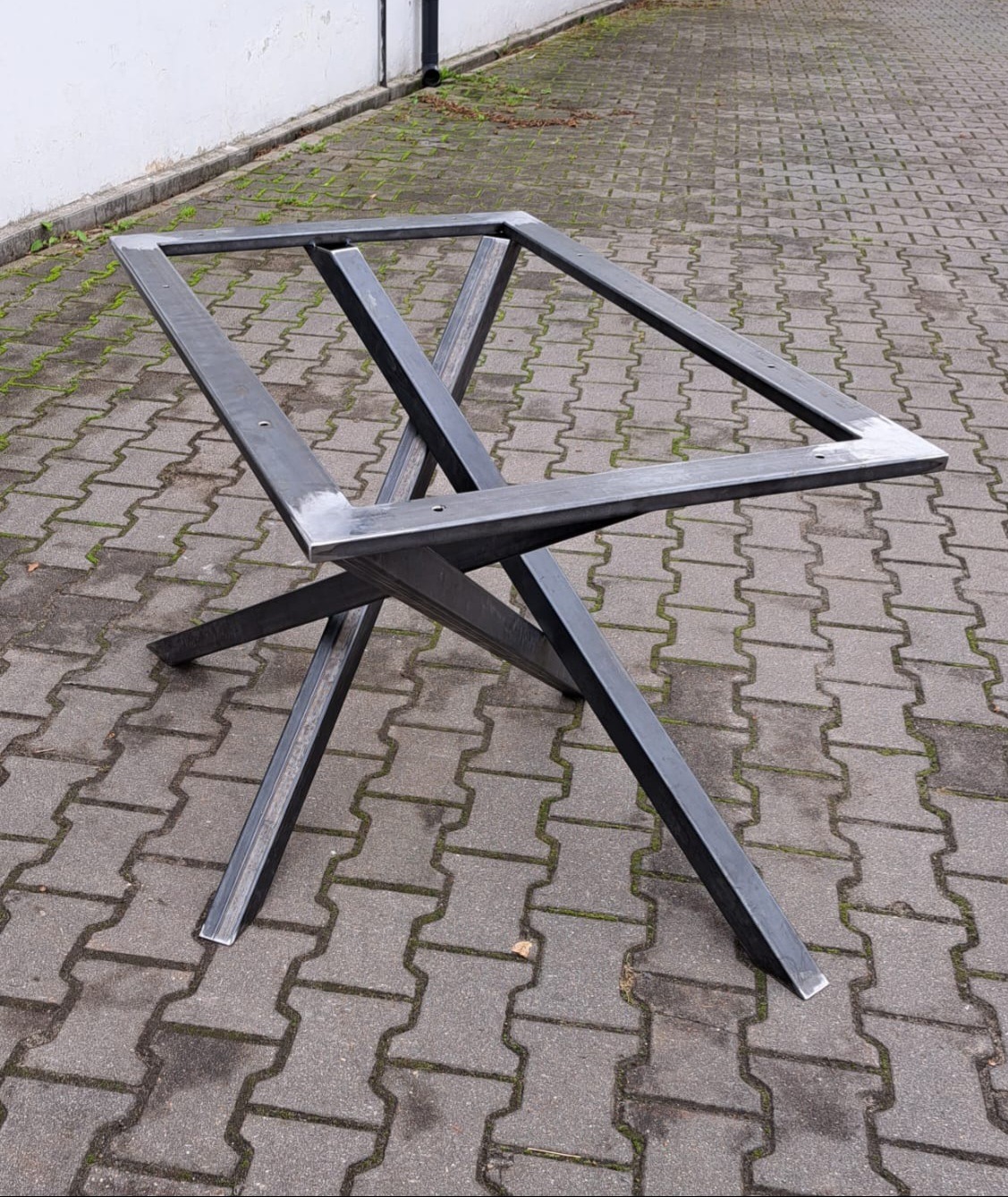 Surowa, spawana rama stołu z profili stalowych, z widocznymi śladami po spawaniu i otworami montażowymi, ustawiona na kostce brukowej.