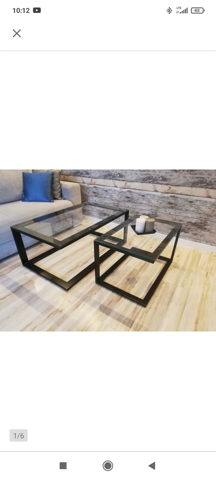 Dwa stoliki kawowe z czarnymi, metalowymi ramami i szklanymi blatami, ustawione na drewnianej podłodze w salonie z szarą sofą i ozdobną ścianą z drewnianych paneli w tle.