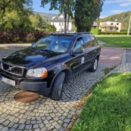 Czarny samochód typu kombi z napisem TAXI na dachu, zaparkowany na brukowanej nawierzchni z widokiem na budynki i zalesione wzgórza w tle.