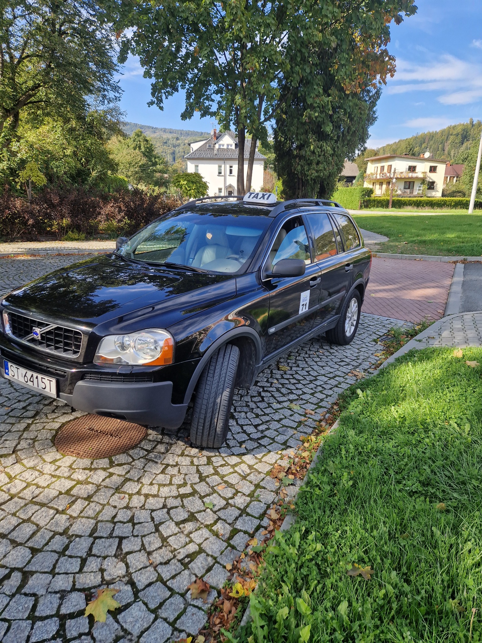 Czarny samochód typu kombi z napisem TAXI na dachu, zaparkowany na brukowanej nawierzchni z widokiem na budynki i zalesione wzgórza w tle.