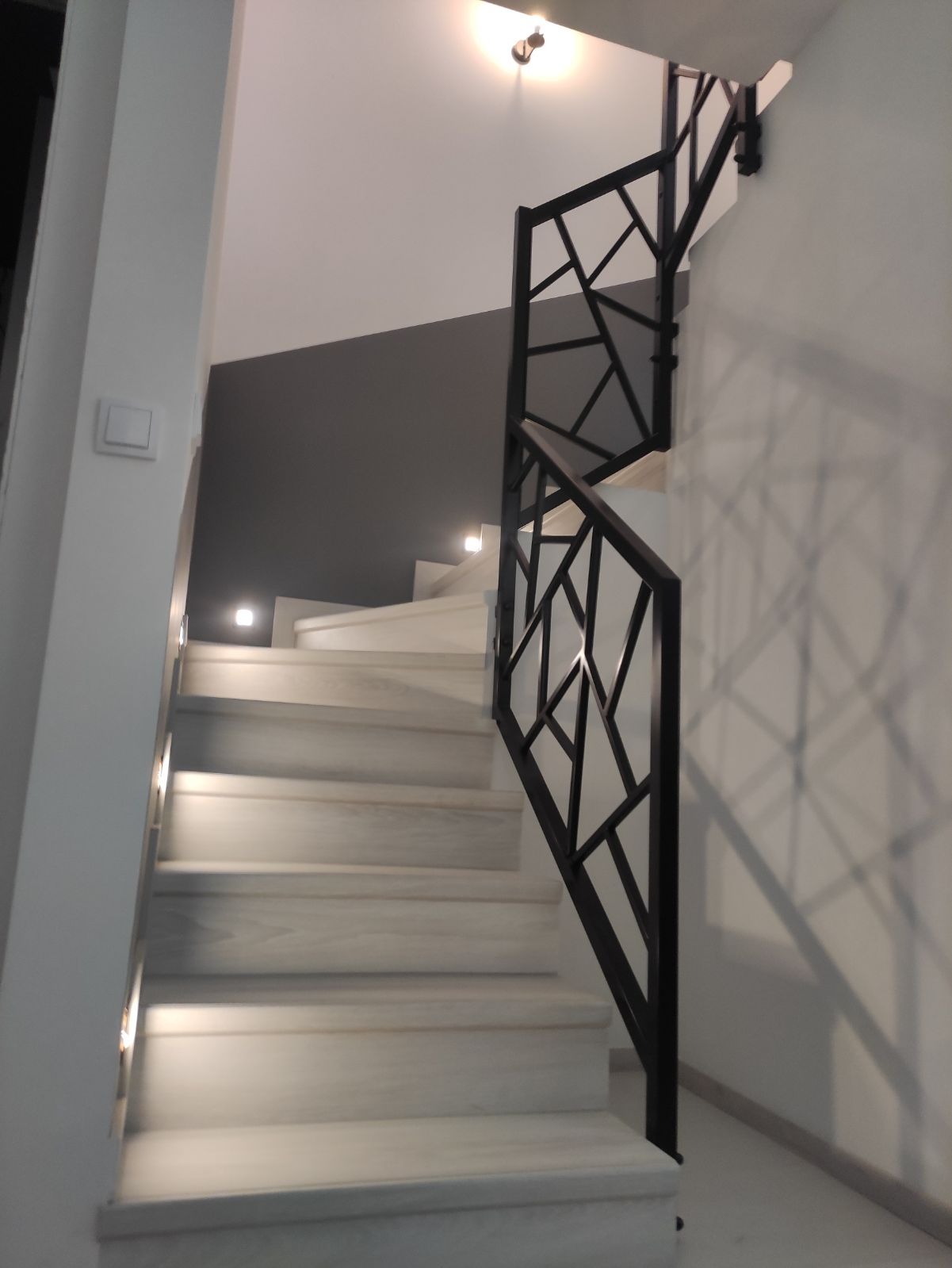 Nowoczesne schody z białymi stopniami i czarną, geometryczną balustradą, oświetlone punktowymi lampkami w ścianie.