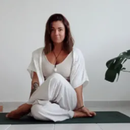 Kobieta w białym stroju siedzi w pozycji lotosu na zielonej macie do ćwiczeń, obok poduszki do medytacji i roślin doniczkowych w jasnym wnętrzu.
