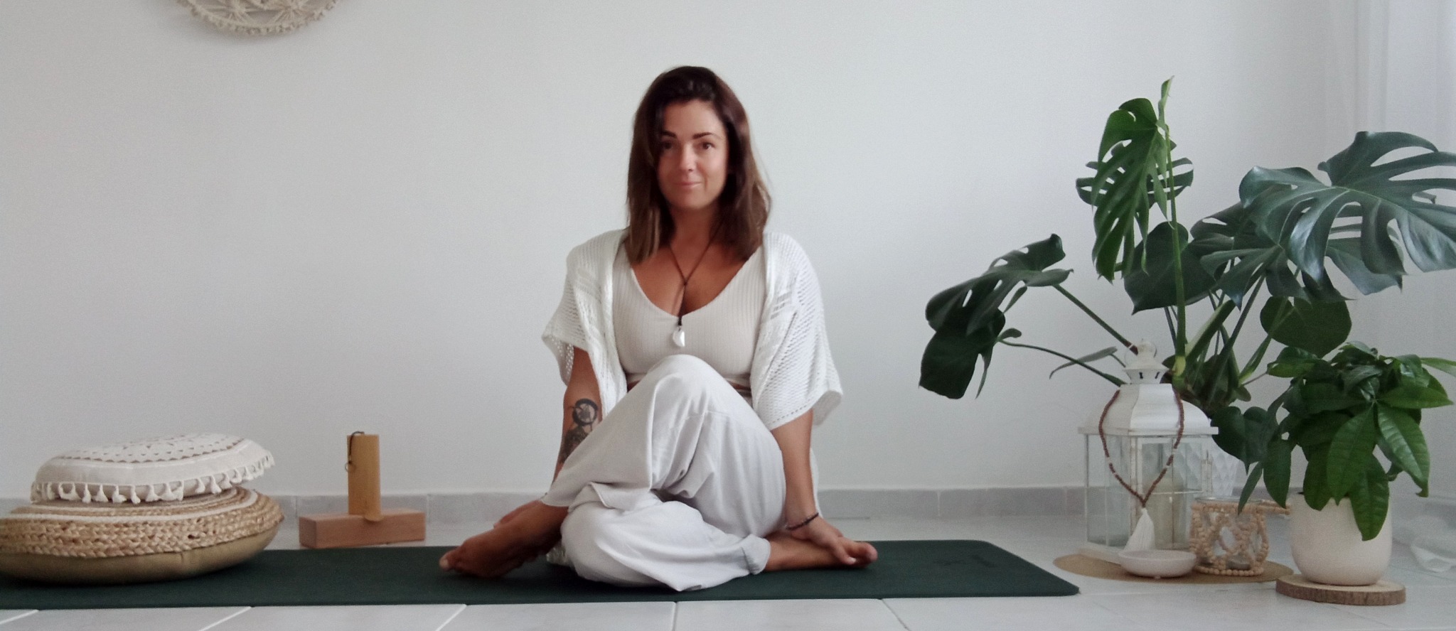 Kobieta w białym stroju siedzi w pozycji lotosu na zielonej macie do ćwiczeń, obok poduszki do medytacji i roślin doniczkowych w jasnym wnętrzu.