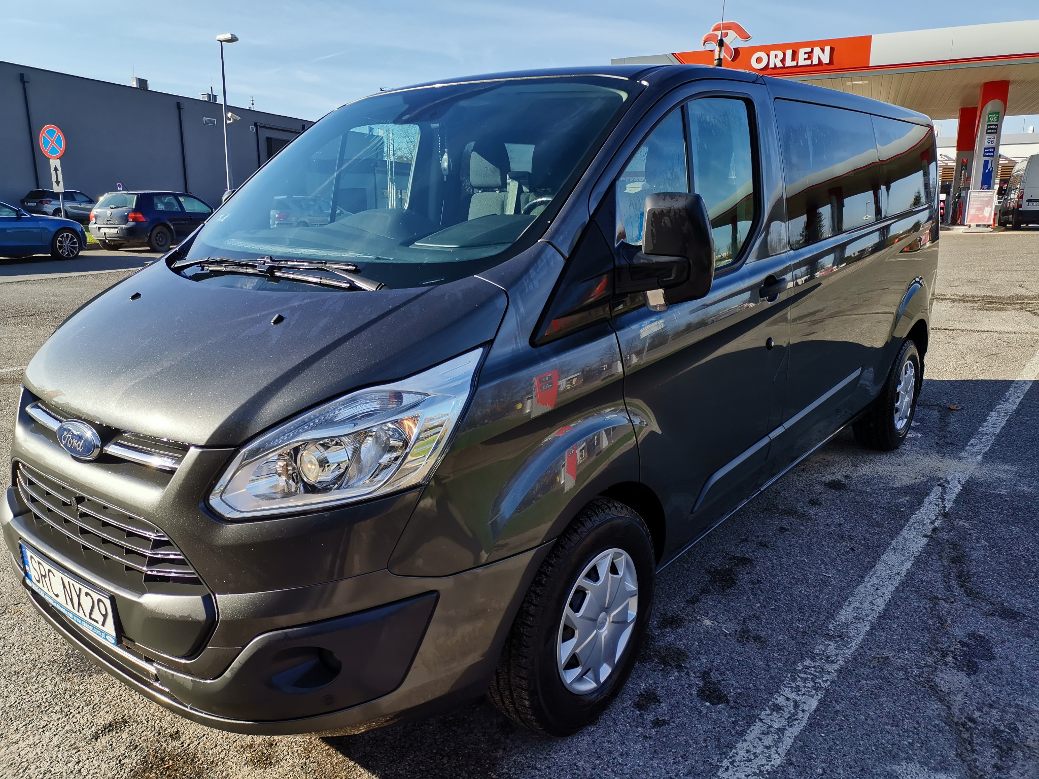 Szary Ford Transit, widok z przodu, zaparkowany przy stacji Orlen, z widoczną tablicą rejestracyjną SRC NYX29.