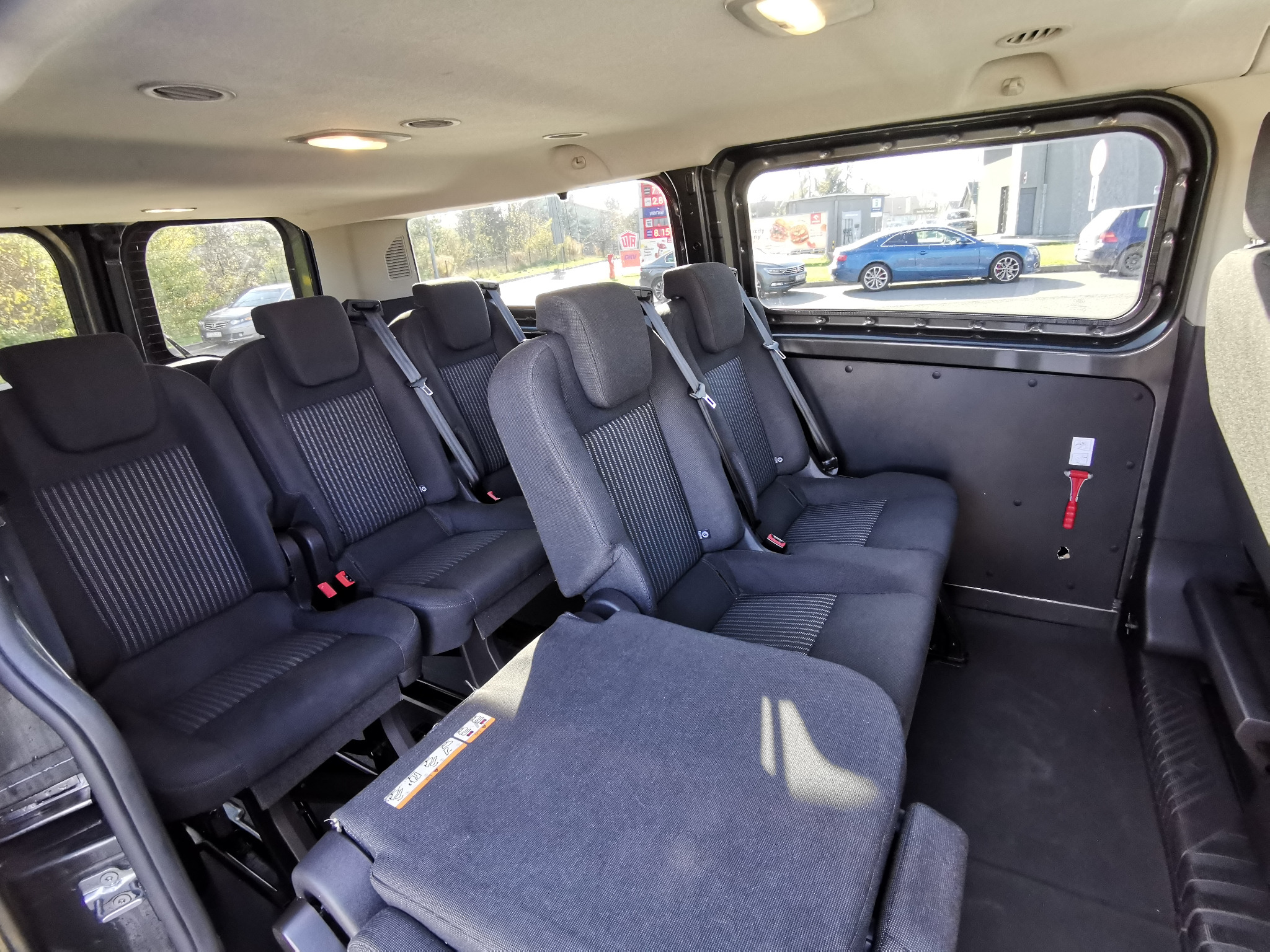 Wnętrze busa z trzema rzędami foteli o ciemnej tapicerce w paski, widok z tyłu, przez okno widoczny niebieski samochód na parkingu.