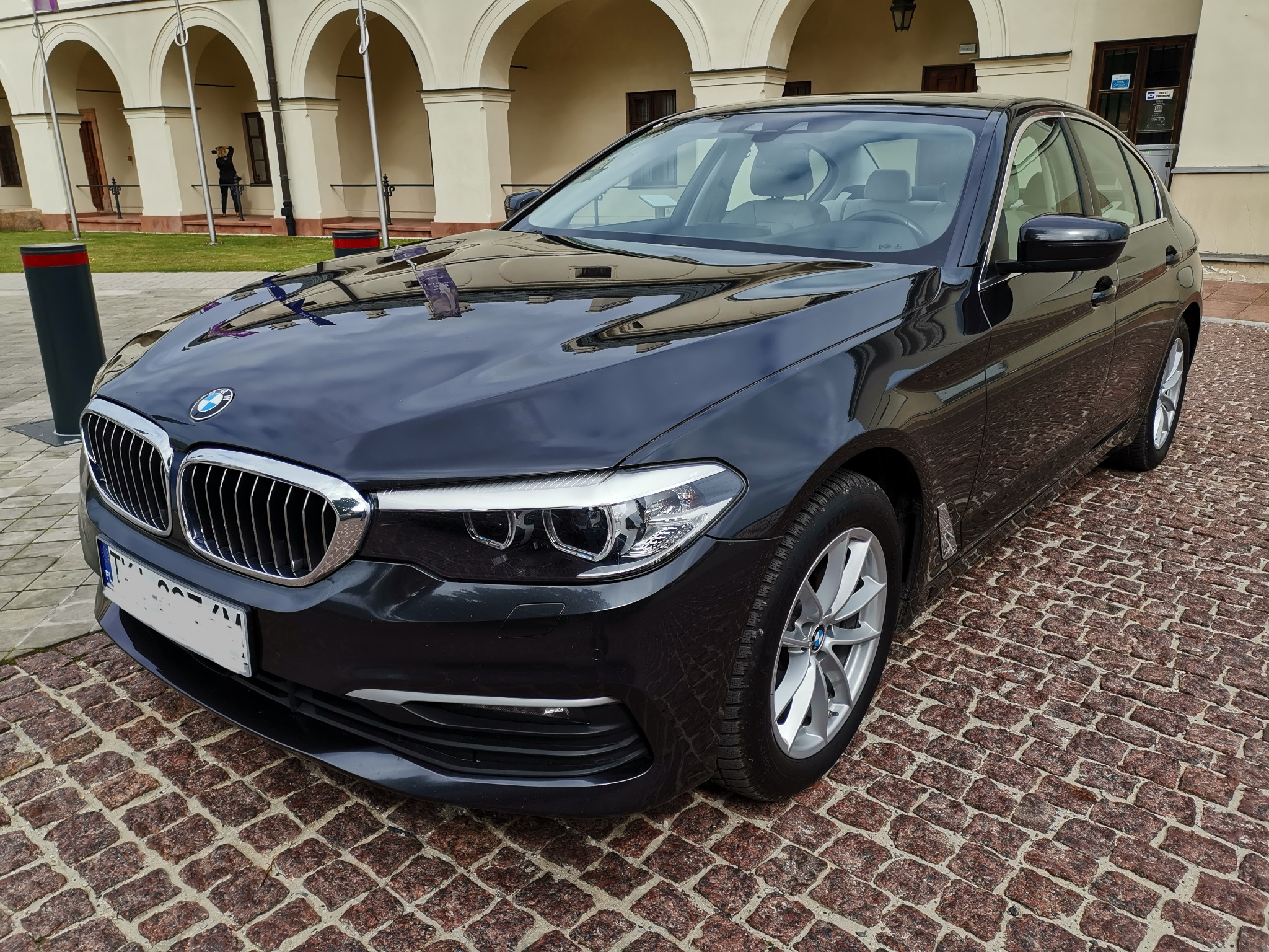 Czarny samochód BMW serii 5 zaparkowany na brukowanej nawierzchni, z budynkiem z arkadami w tle i osobą robiącą zdjęcie w oddali.