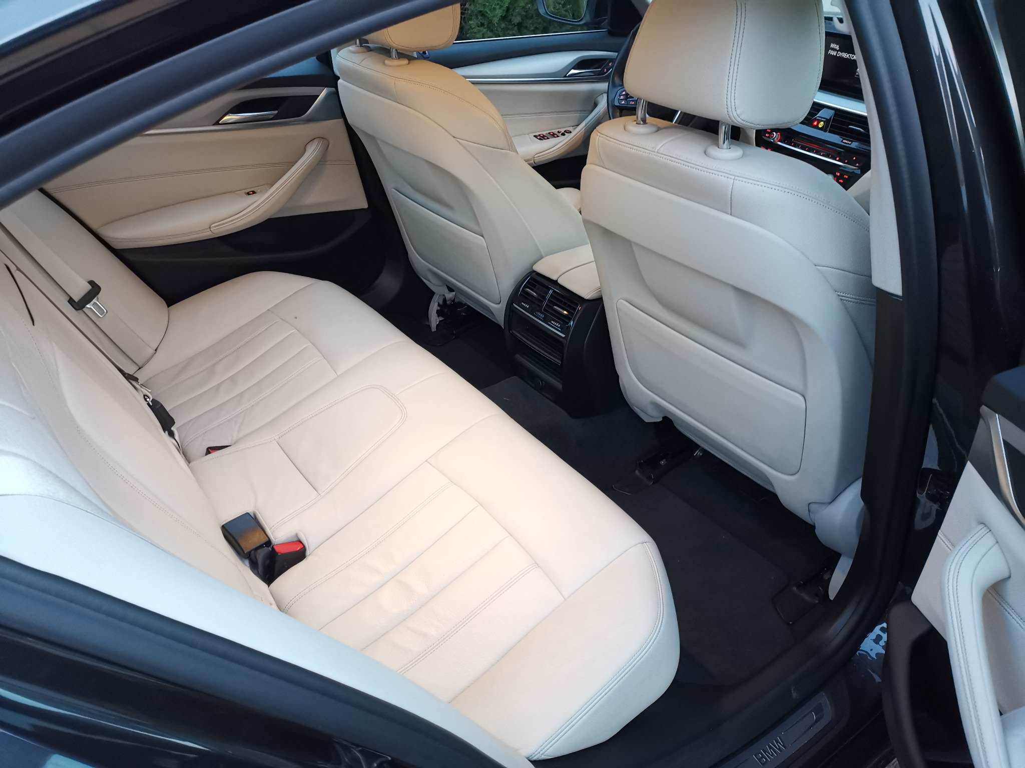 Wnętrze samochodu z jasną skórzaną tapicerką, widok na tylną kanapę z pasami bezpieczeństwa i zagłówkami, czarna podłoga, elementy deski rozdzielczej w tle.