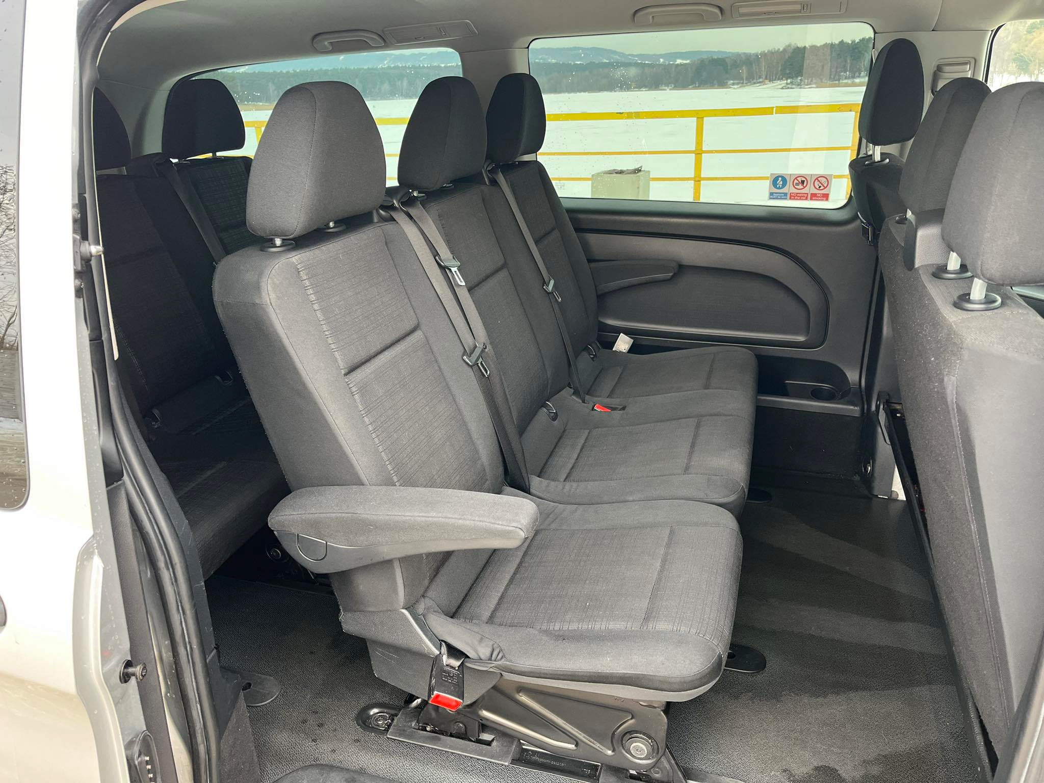 Wnętrze busa z trzema rzędami ciemnych, materiałowych siedzeń, widok z otwartych drzwi, za oknem zimowy krajobraz z jeziorem i żółtą barierką.