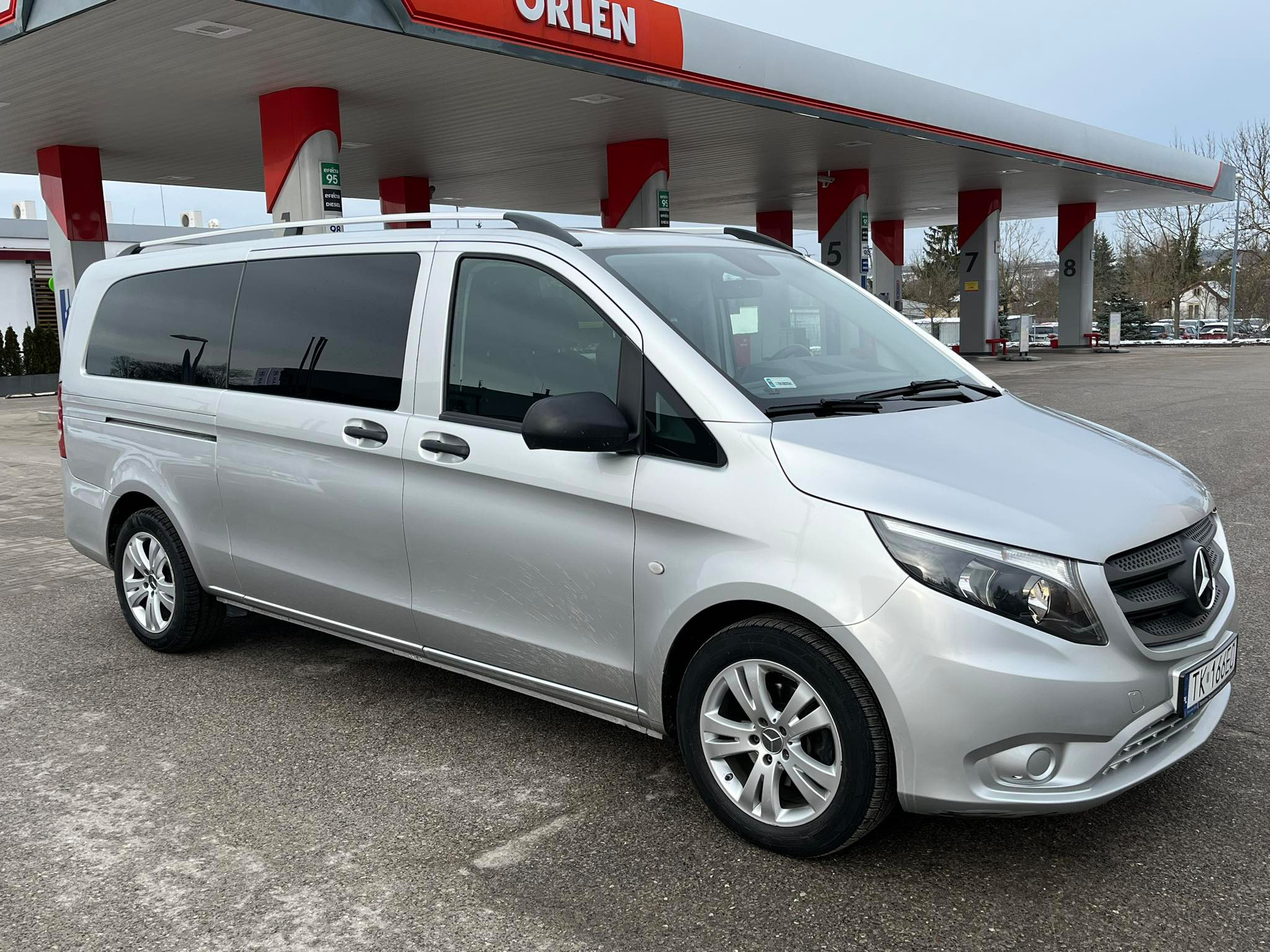 Srebrny van Mercedes-Benz Vito z relingami dachowymi zaparkowany na stacji benzynowej Orlen w pochmurny dzień.