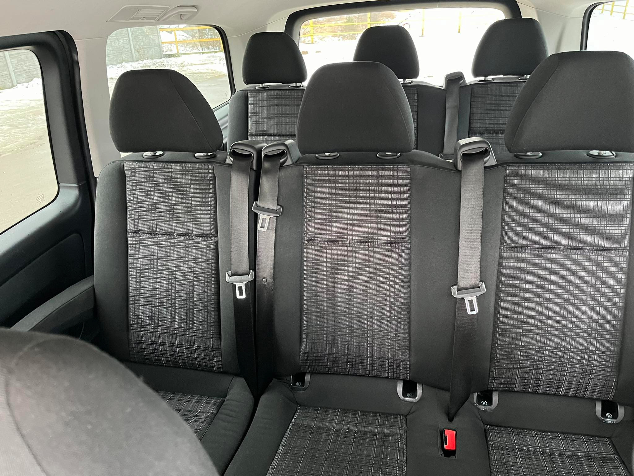 Wnętrze busa z trzema rzędami foteli z tapicerką w drobną szarą kratkę i pasami bezpieczeństwa, widok z tyłu.