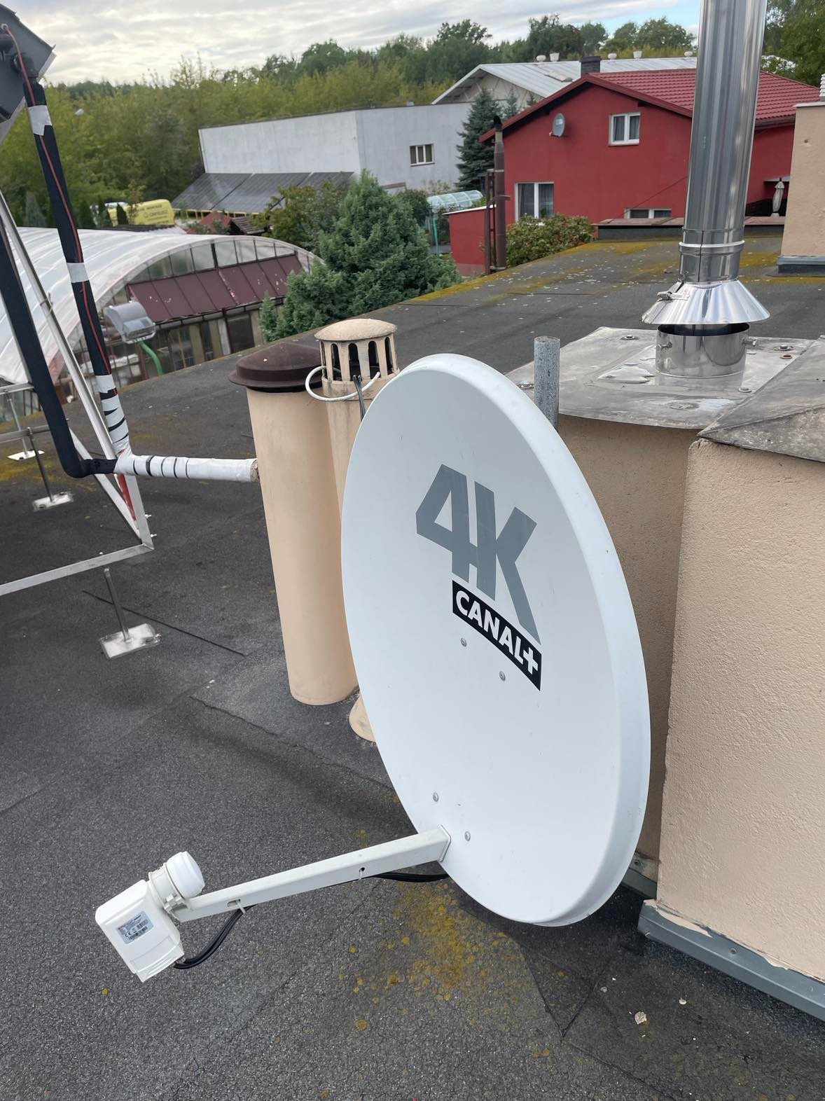 Biała antena satelitarna 4K Canal+ zamontowana na płaskim, bitumicznym dachu, widoczny komin i inne instalacje dachowe, w tle zabudowa jednorodzinna.