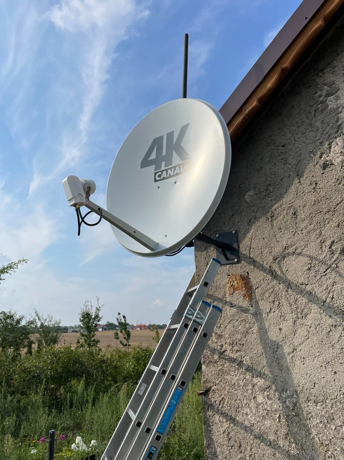 Antena satelitarna 4K z logo Canal+ zamontowana na szarej ścianie domu w Brodnicy, widoczna drabina oparta o ścianę, tło stanowi błękitne niebo i zieleń.