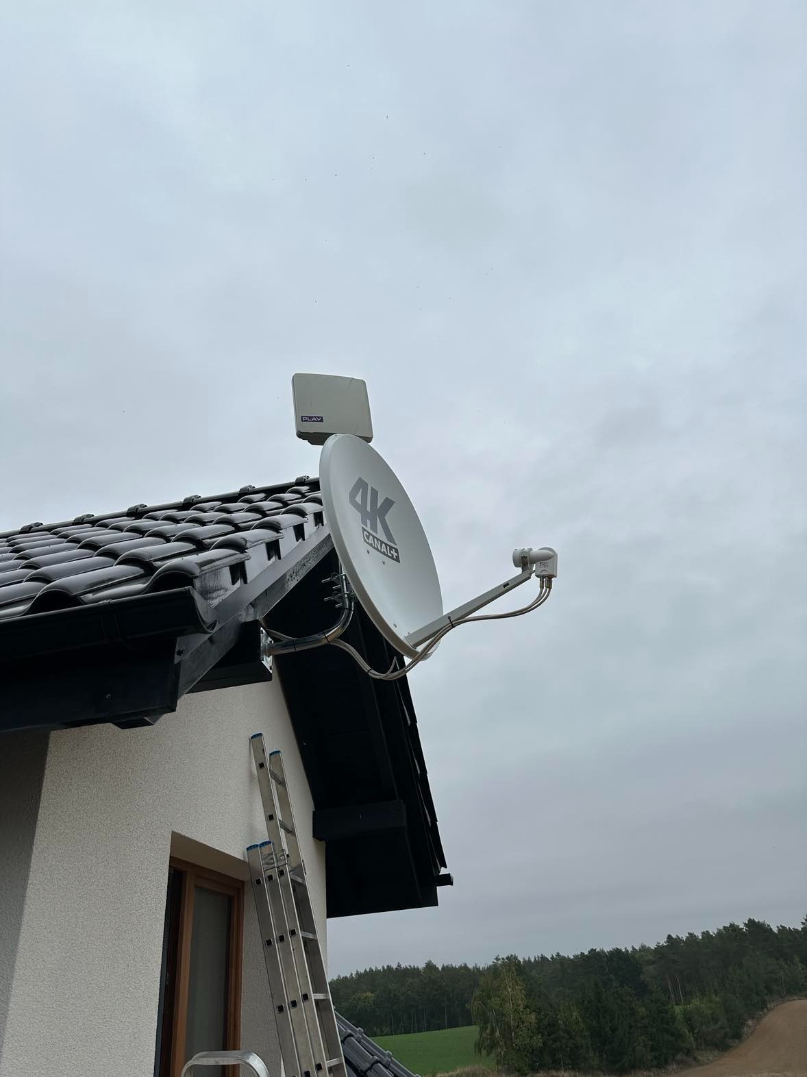 Antena satelitarna 4K Canal+ i antena Play zamontowane na dachu domu jednorodzinnego z czarną dachówką, drabina oparta o ścianę, pochmurne niebo w tle.