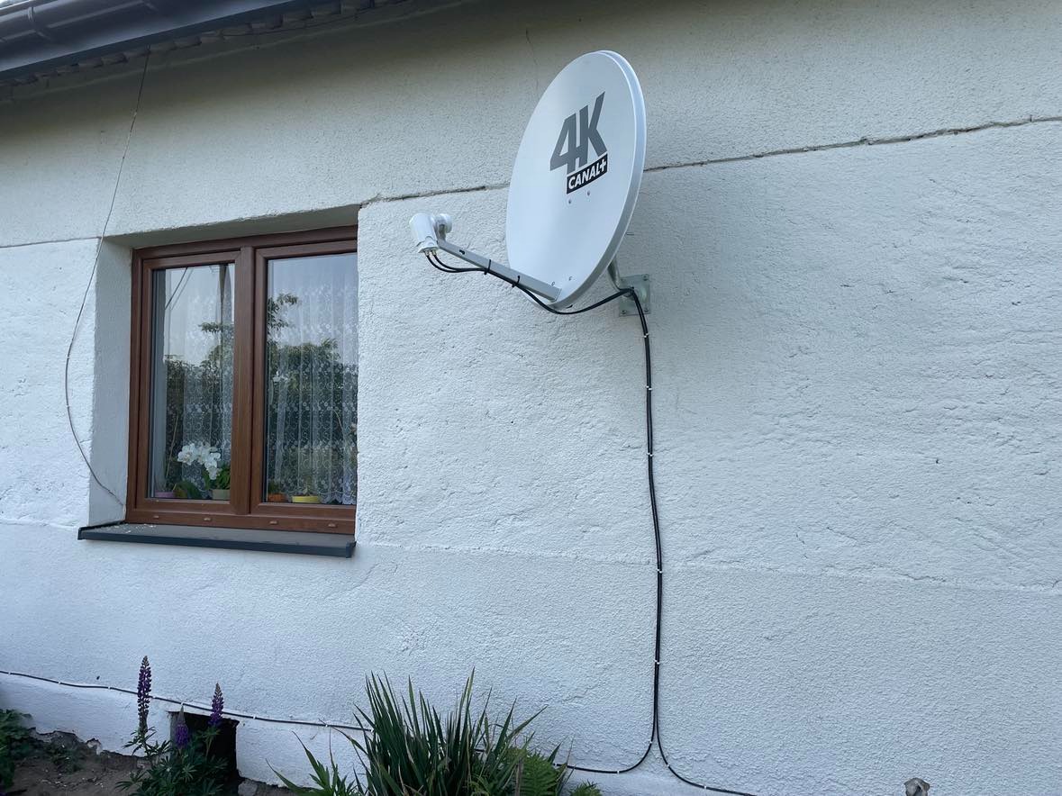 Biała antena satelitarna 4K Canal+ zamontowana na elewacji domu obok okna z brązową ramą, kabel przymocowany do ściany klamrami, widoczne rośliny przy fundamencie.