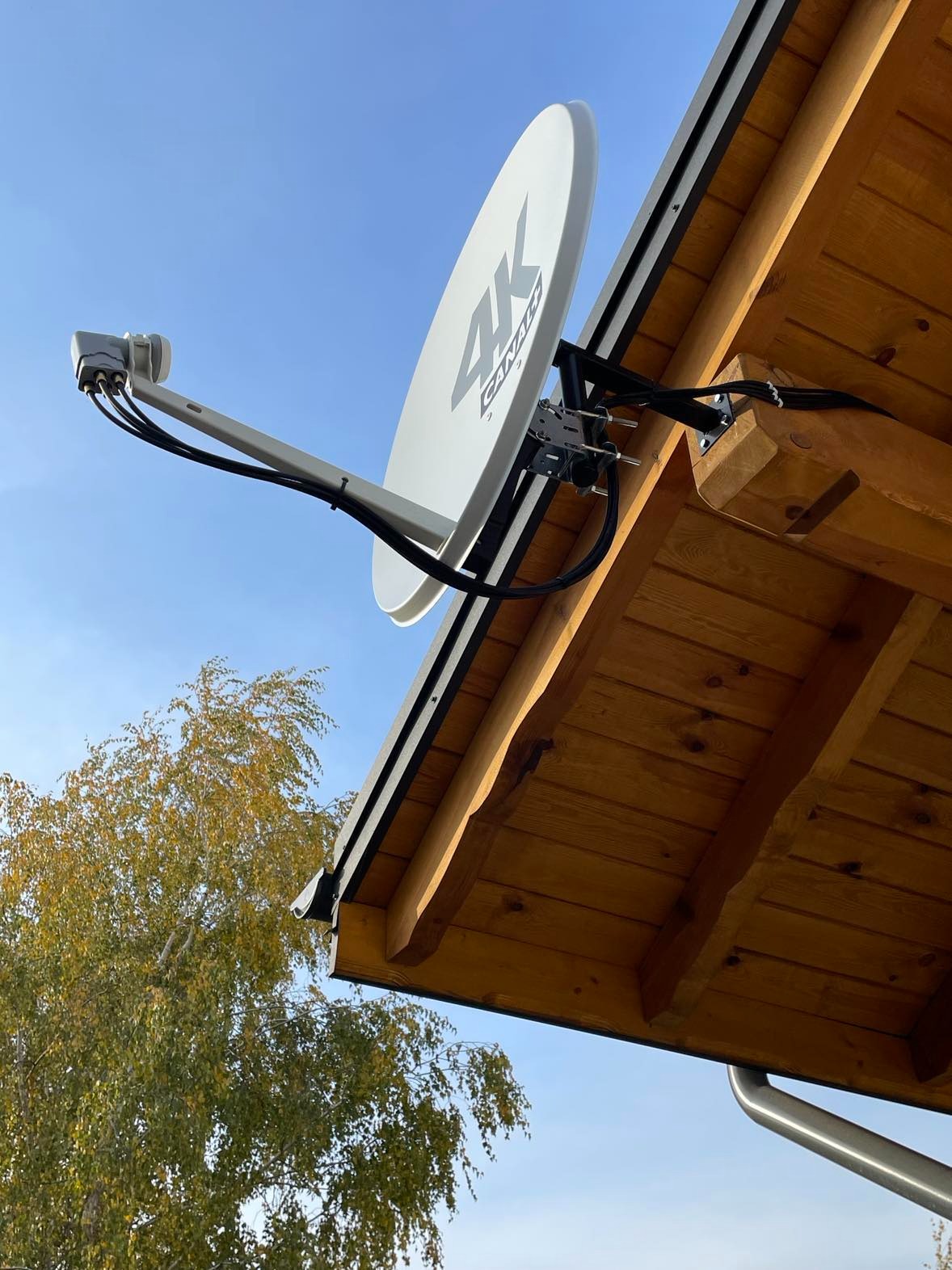 Biała antena satelitarna z logo 4K Canal+ zamontowana na drewnianym okapie dachu, widok z dołu na tle błękitnego nieba i jesiennego drzewa.