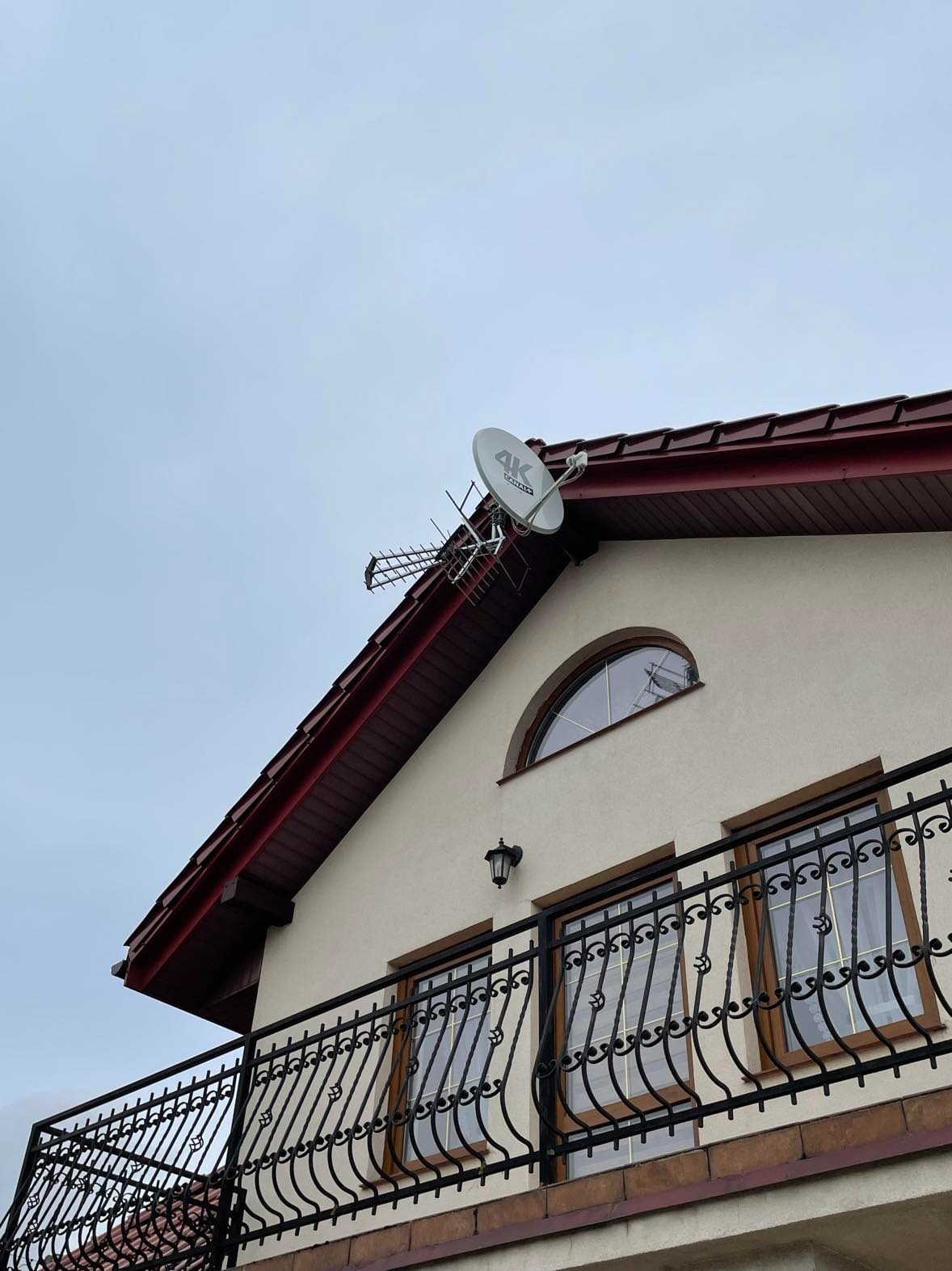 Antena satelitarna 4K i antena naziemna zamontowane na dachu domu jednorodzinnego z balkonem, widok z dołu na pochmurne niebo.
