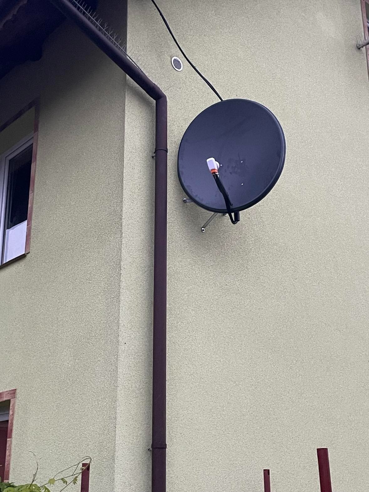 Czarna antena satelitarna zamontowana na elewacji budynku o jasnej, piaskowej barwie, widoczny brązowy przewód odprowadzający wodę deszczową oraz fragment okna.