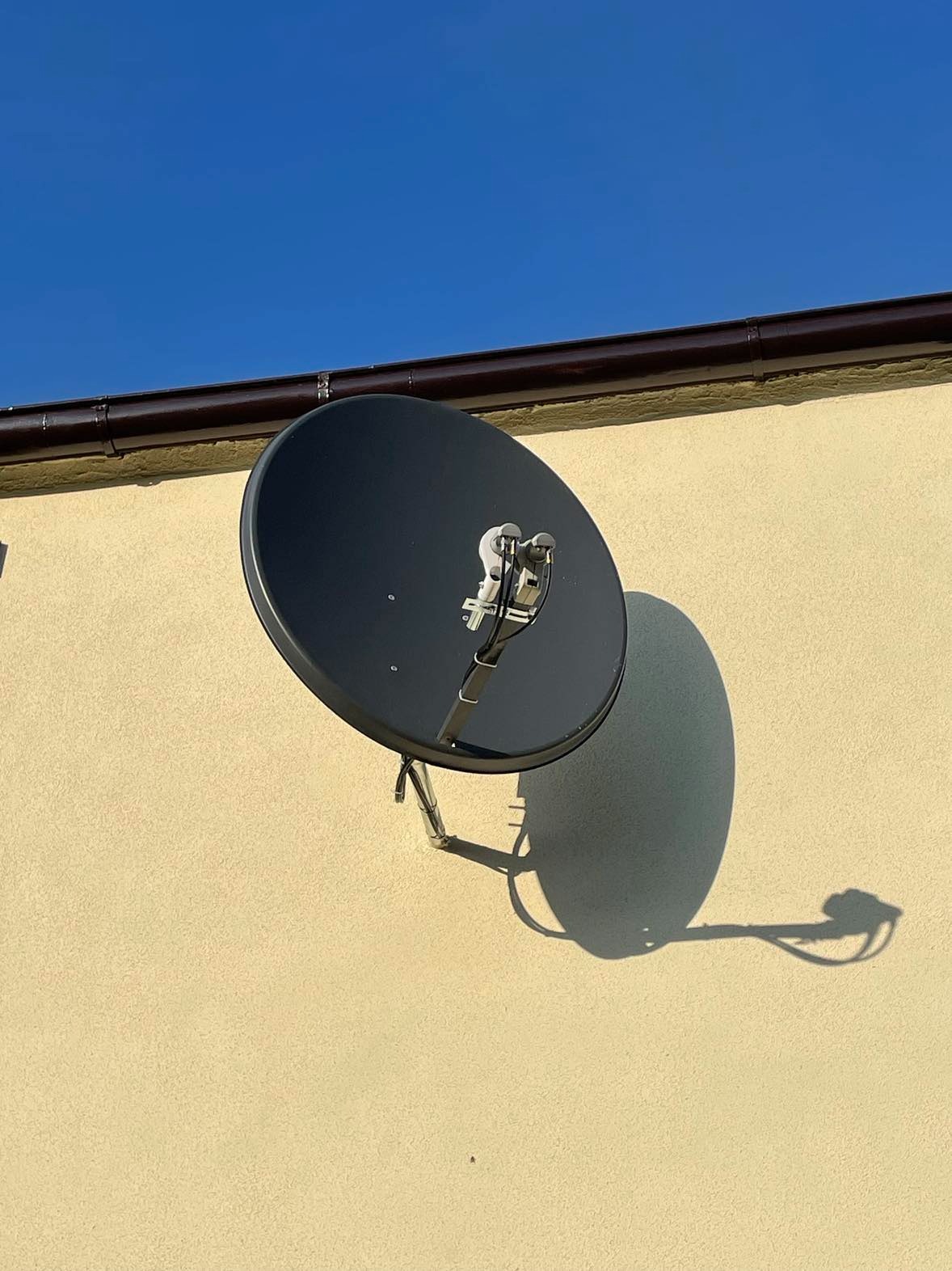 Czarna antena satelitarna zamontowana na beżowej ścianie domu z widoczną rynną na tle błękitnego nieba, rzucająca wyraźny cień w słoneczny dzień.