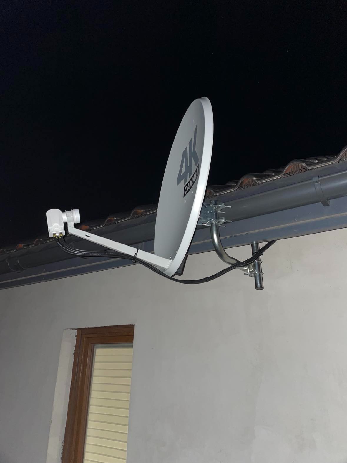 Biała antena satelitarna 4K Canal+ zamontowana na dachu budynku, widoczna instalacja kablowa i uchwyty mocujące, tło stanowi ciemne nocne niebo.