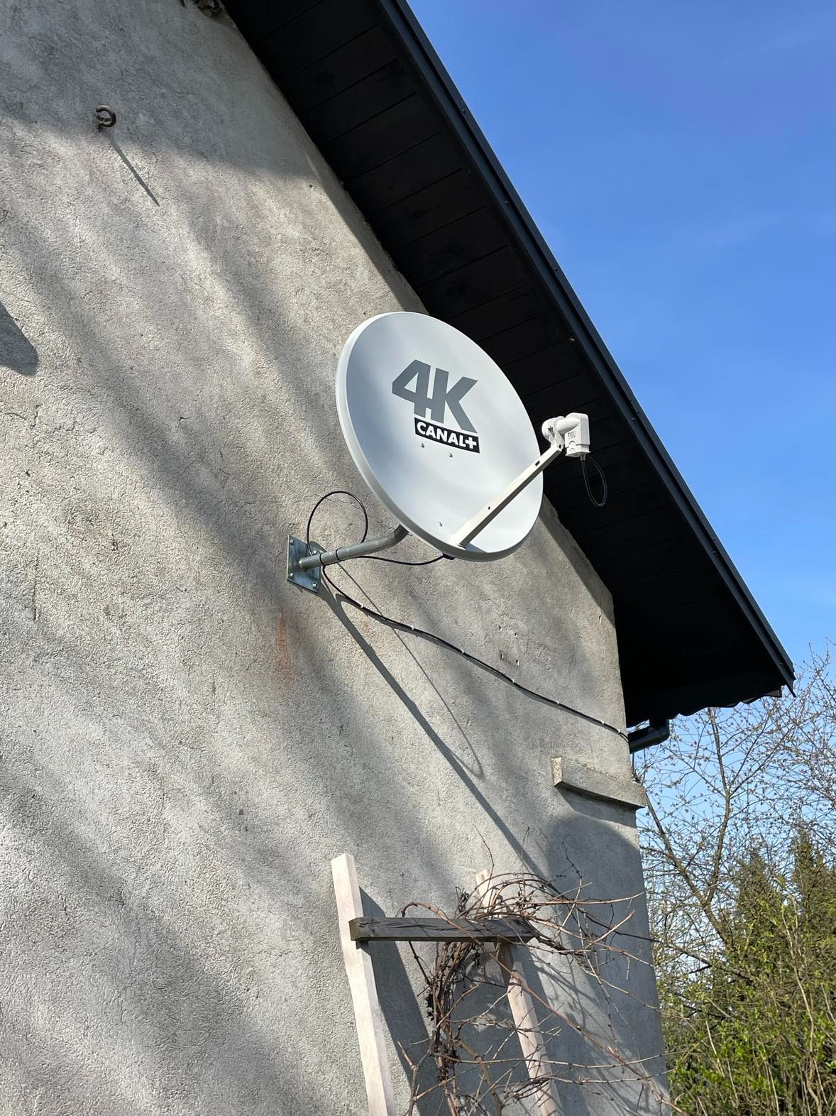 Biała antena satelitarna 4K Canal+ zamontowana na szarej, chropowatej ścianie budynku, widoczna z dołu pod kątem, z fragmentem dachu i niebem w tle.