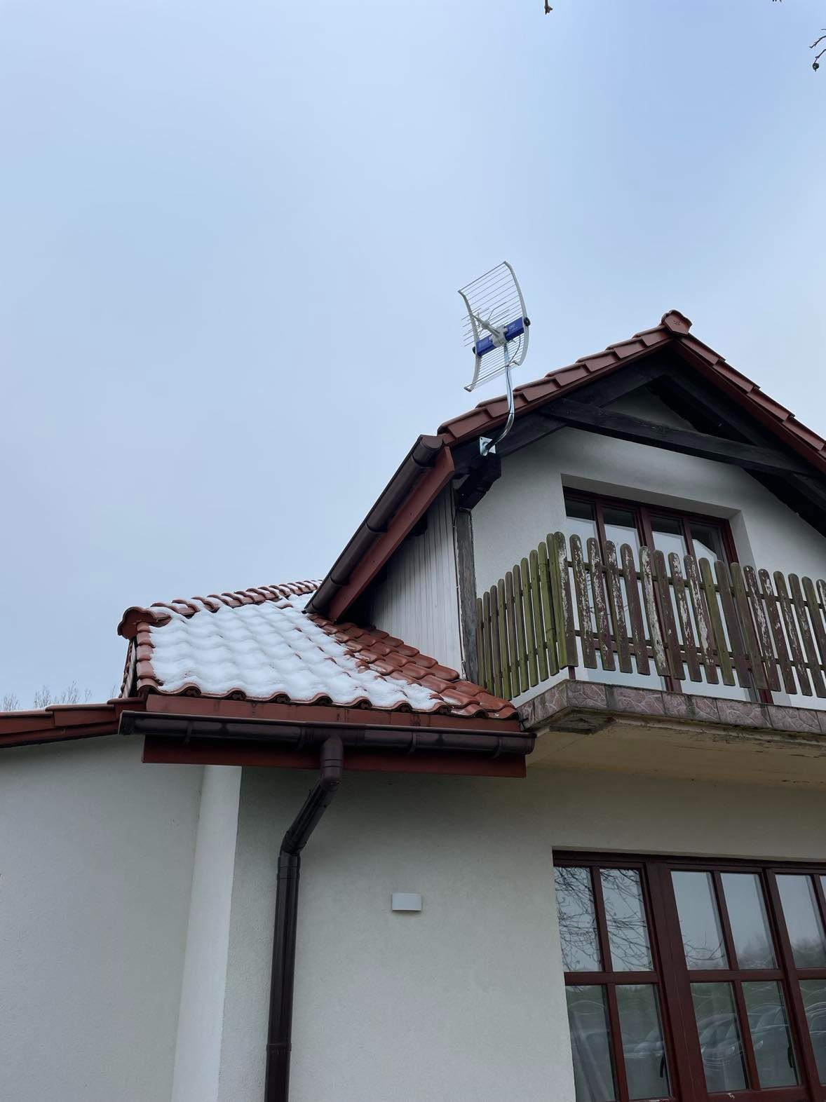 Antena telewizyjna typu Yagi-Uda zamontowana na dachu domu jednorodzinnego pokrytego czerwoną dachówką z resztkami śniegu, widoczna z dołu na tle pochmurnego nieba.