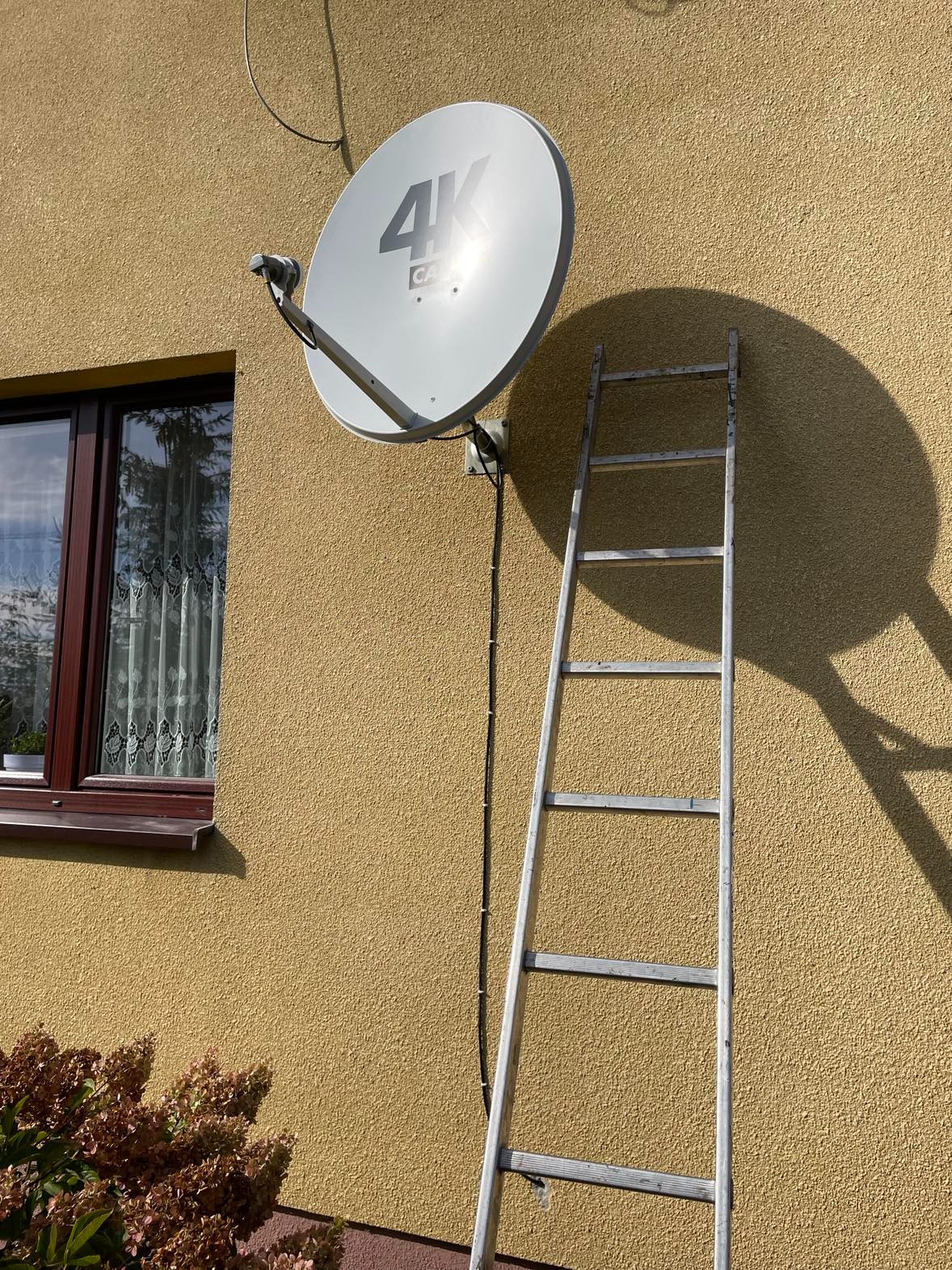 Antena satelitarna 4K zamontowana na żółtej elewacji domu, obok okna z brązową ramą i drabiny oparta o ścianę, cień anteny na ścianie.