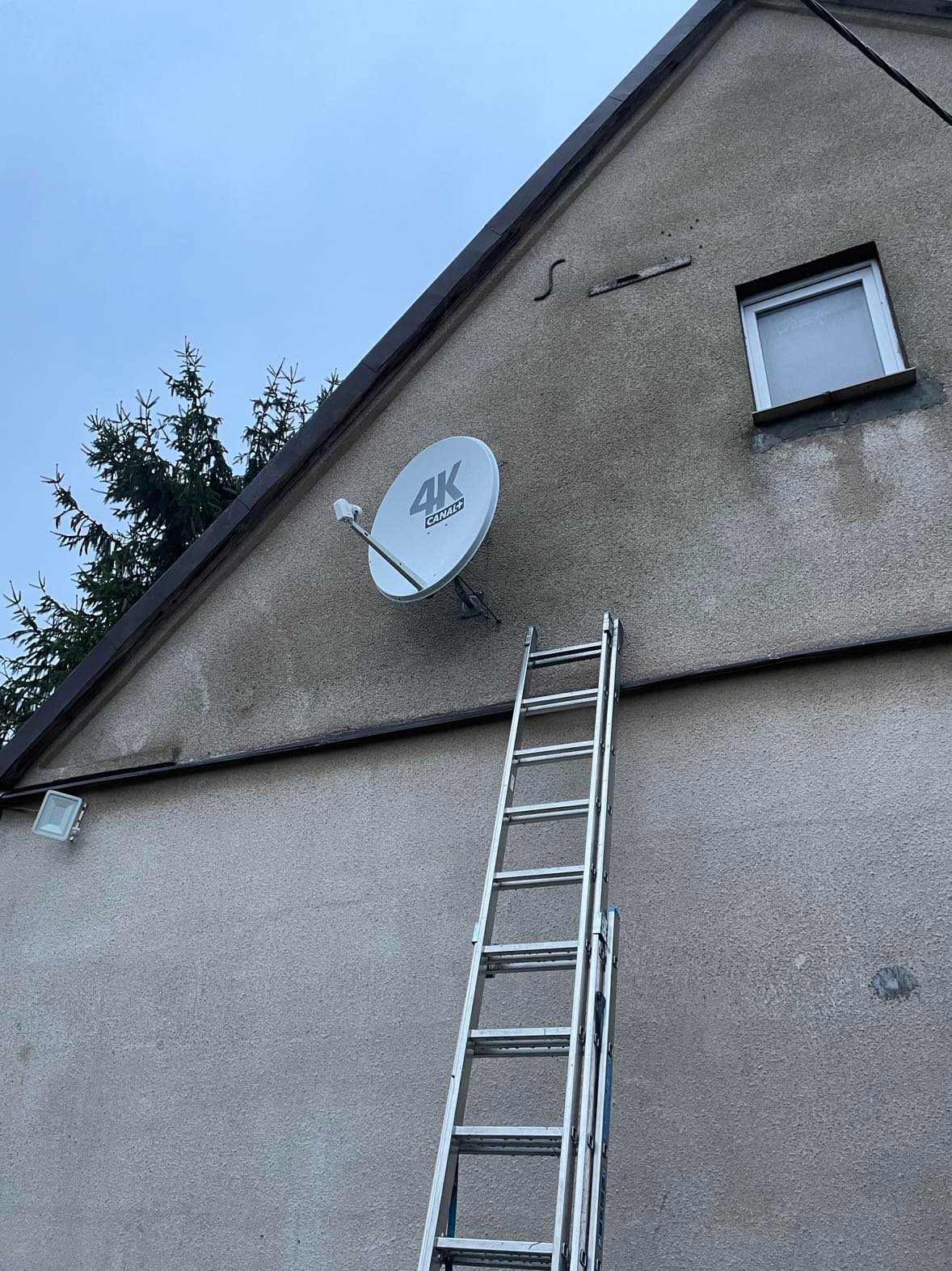 Antena satelitarna 4K Canal+ zamontowana na szczycie domu z pomocą aluminiowej drabiny, widok z dołu na tle pochmurnego nieba.