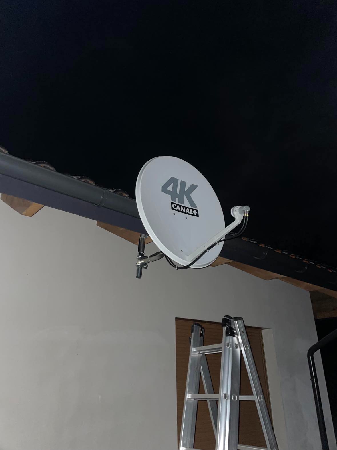 Biała antena satelitarna 4K Canal+ zamontowana na ścianie domu pod okapem dachu, z widoczną drabiną opartą o ścianę.