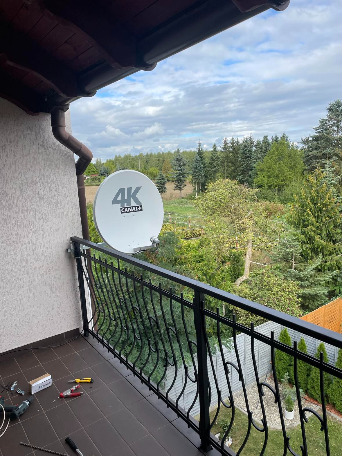 Biała antena satelitarna 4K Canal+ zamontowana na balkonie z czarną, metalową balustradą, widok na zalesiony teren, narzędzia pracy rozłożone na podłodze balkonu.