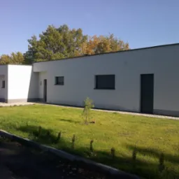 Nowoczesny, parterowy dom z białą elewacją i płaskim dachem, otoczony trawnikiem i młodymi drzewkami iglastymi.