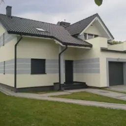 Dwukondygnacyjny dom jednorodzinny z garażem w bryle, kremowa elewacja z szarymi pasami, ciemna dachówka i stolarka okienna, widok z poziomu trawnika.