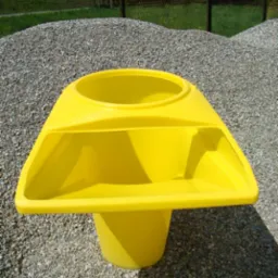 Żółty zsyp do gruzu z tworzywa sztucznego, ustawiony na żwirowym podłożu, z widocznym w tle kopcem żwiru i tablicą informacyjną.