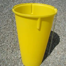 Żółty, plastikowy zsyp do gruzu z metalowym łańcuchem, stojący na żwirowym podłożu z widoczną górą żwiru w tle.