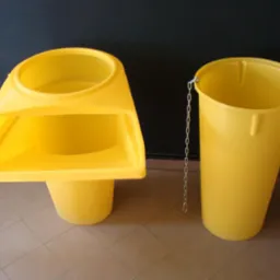 Dwa żółte zsypy do gruzu: jeden kompletny z lejkiem, drugi w formie wiadra z łańcuchem, na tle szaro-beżowej podłogi i ciemnej ściany.