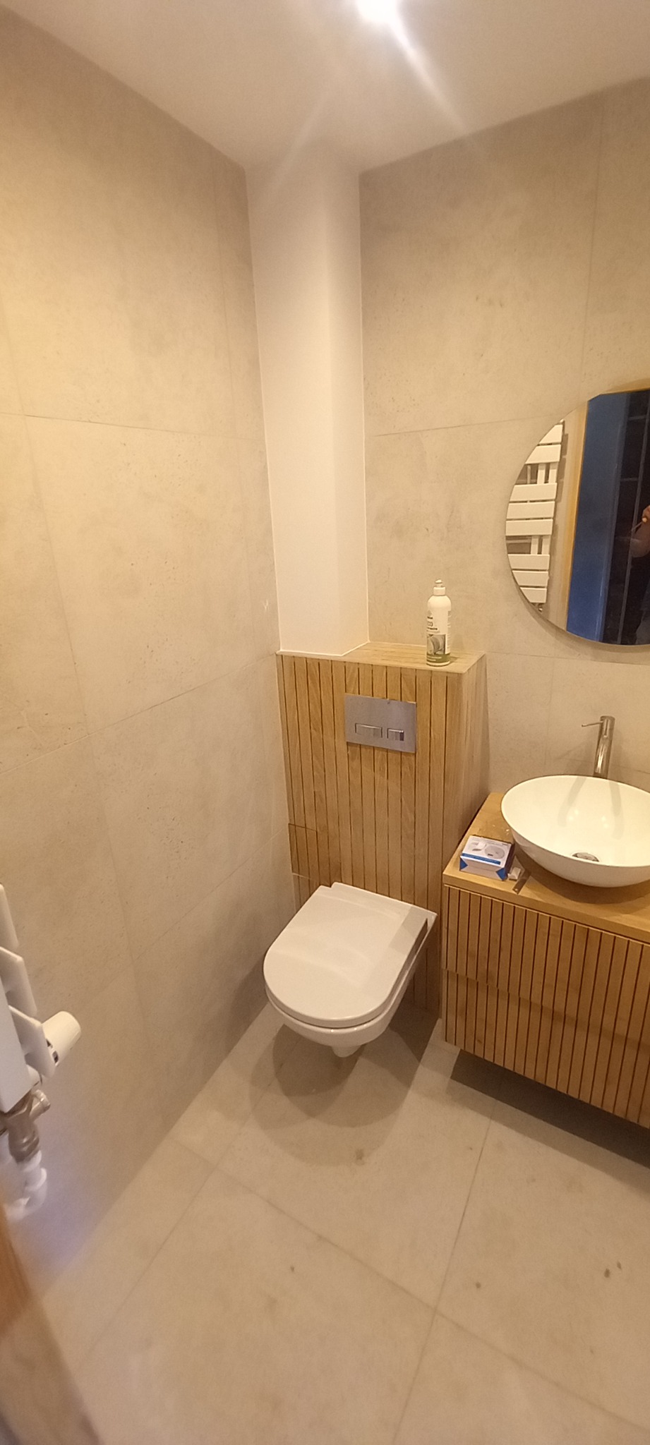 Jasna łazienka z drewnianymi akcentami: obudowa WC, szafka pod umywalkę z okrągłym lustrem. Minimalistyczny design, beżowe płytki na ścianach i podłodze.