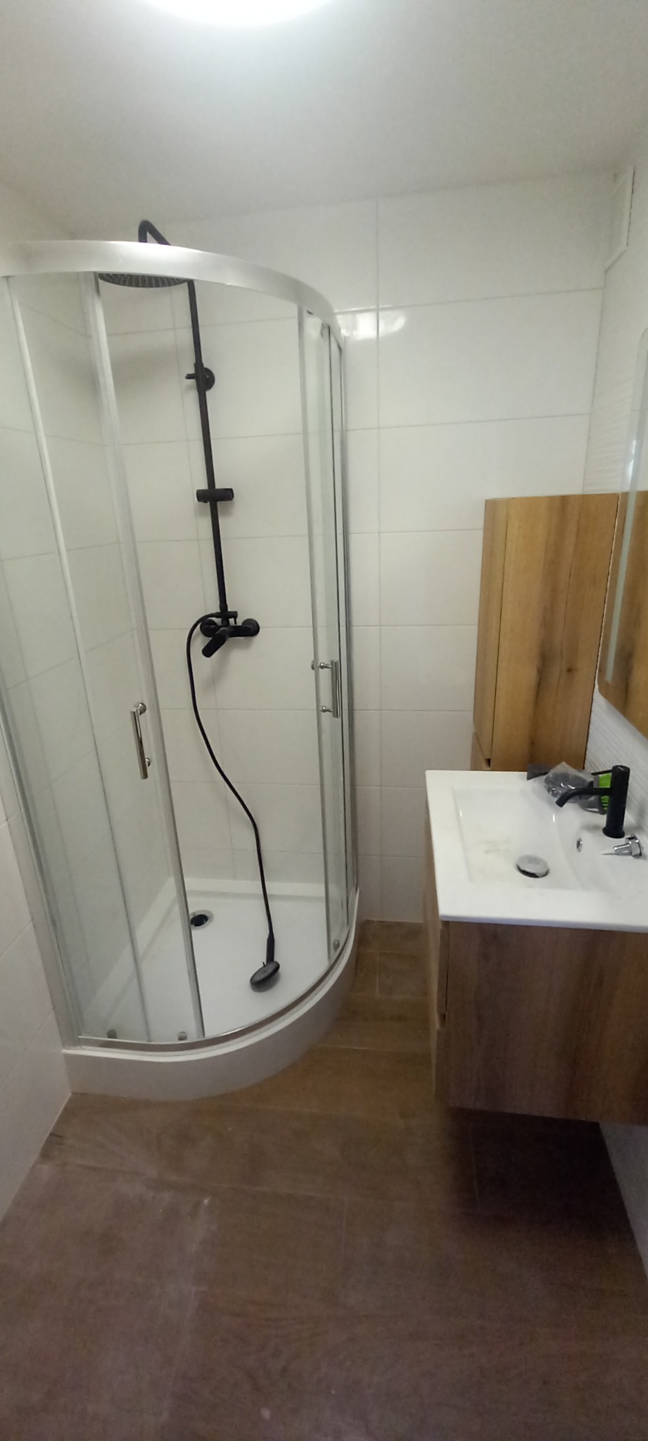 Nowoczesna łazienka po remoncie: kabina prysznicowa z czarną armaturą, umywalka z szafką w kolorze drewna, białe płytki ścienne i brązowe płytki podłogowe.