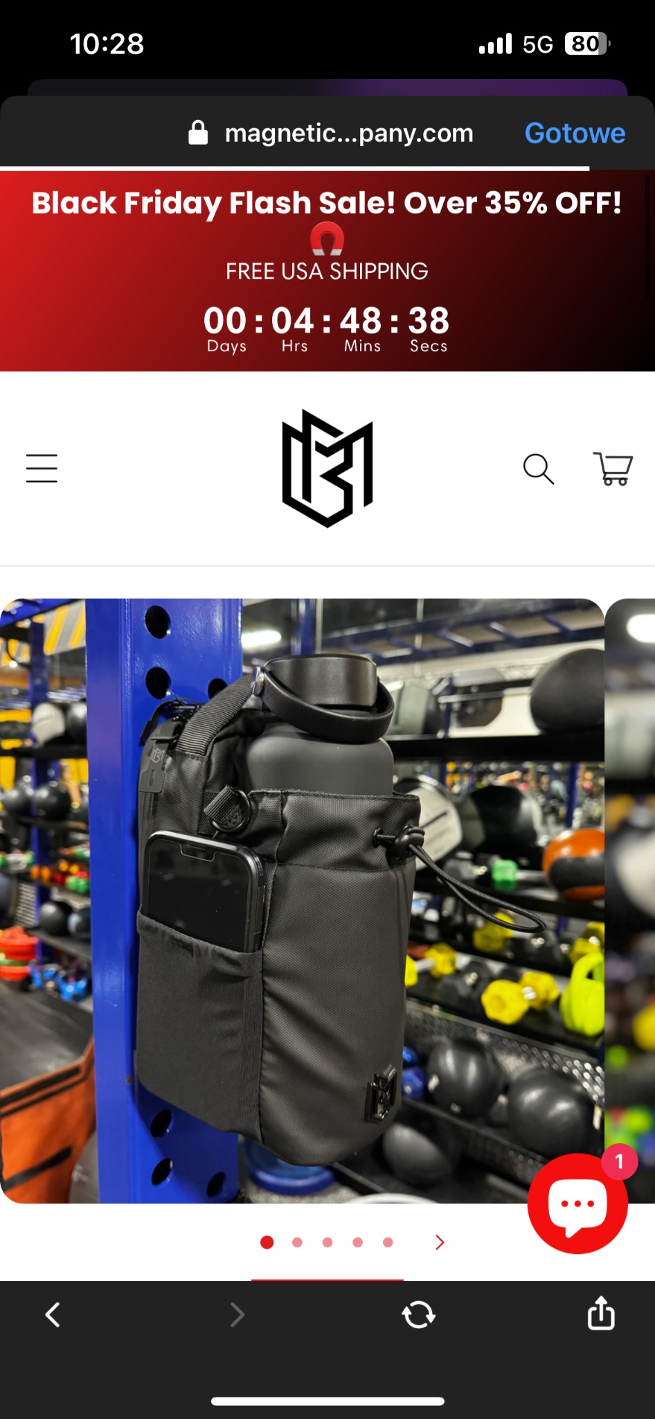 Zdjęcie z ekranu telefonu prezentujące czarny bidon w torbie z kieszenią na telefon, zawieszone na niebieskim stojaku na tle sprzętu do ćwiczeń. Na górze ekranu widoczny baner reklamowy Black Friday.