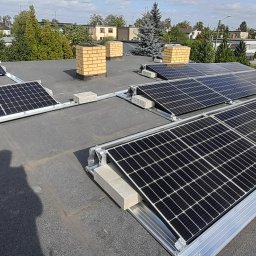 TO GLORY Oleh Blahodyr - Panele słoneczne zamontowane na płaskim dachu budynku z widocznymi kominami i cieniem osoby robiącej zdjęcie.