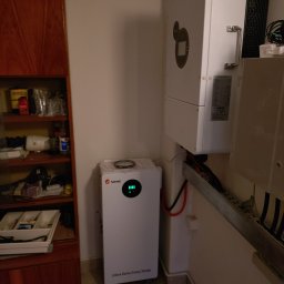 TO GLORY Oleh Blahodyr - Biały magazyn energii SRNE i inwerter na ścianie w pomieszczeniu. Widoczne kable i elementy instalacji. Obok szafka z wyposażeniem.