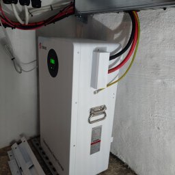 TO GLORY Oleh Blahodyr - Biały magazyn energii SRNE z podłączonymi kablami, zamontowany w rogu pomieszczenia. Widoczne elementy montażowe i kable w peszlach.