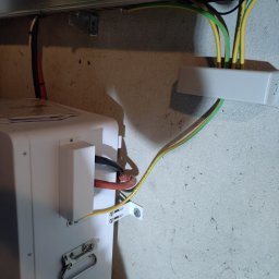 TO GLORY Oleh Blahodyr - Podłączenie przewodów do białego urządzenia, prawdopodobnie magazynu energii, z widocznymi kablami ochronnymi i zasilającymi, zamocowanego na szarej ścianie.