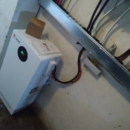 TO GLORY Oleh Blahodyr - Biały magazyn energii SRNE z zielonymi diodami, zamontowany na ścianie, widoczne kable i metalowa szyna. Na górze urządzenia leży kartonowe pudełko.