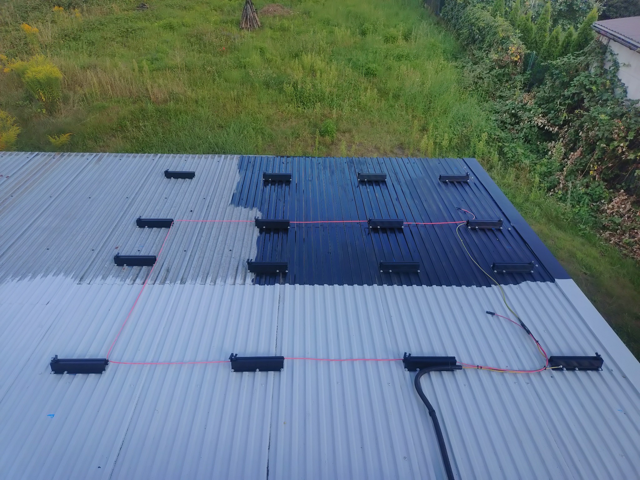 Montaż instalacji fotowoltaicznej na dachu z blachy falistej. Widoczne czarne uchwyty, okablowanie i częściowo pomalowany dach na ciemny kolor. Prace w toku.
