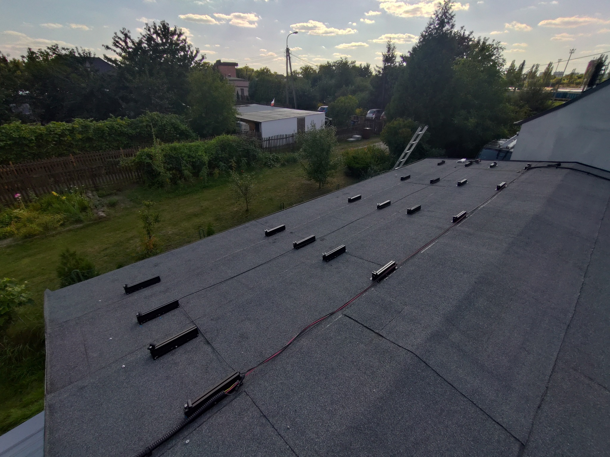 Montaż elementów instalacji fotowoltaicznej na dachu pokrytym papą. Widoczne uchwyty montażowe i przewody. W tle zieleń drzew i zabudowania.