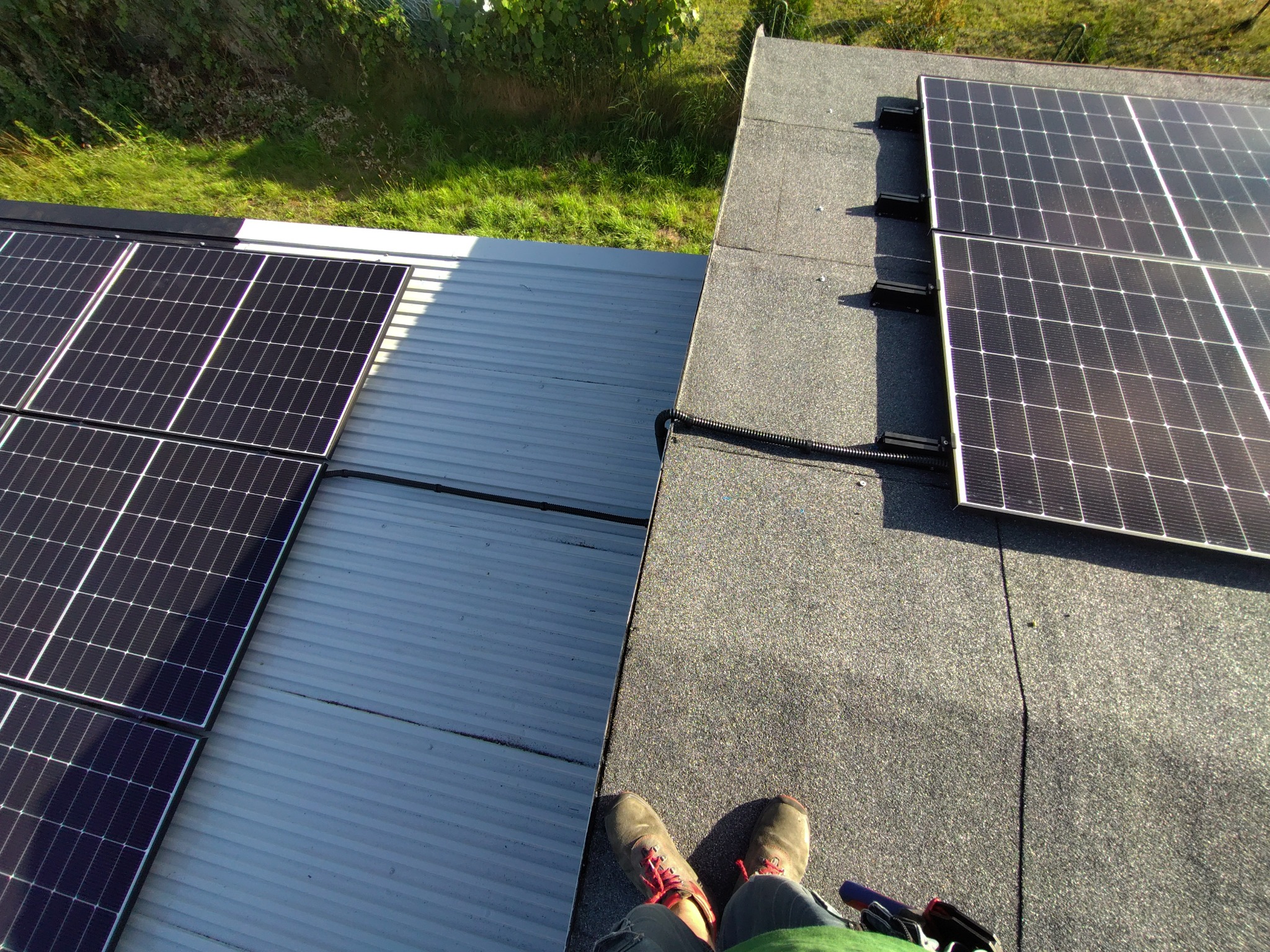 Montaż paneli słonecznych na dachu z papy i blachy falistej. Widok z góry na instalatora w roboczych butach. Częściowo zamontowane panele fotowoltaiczne.