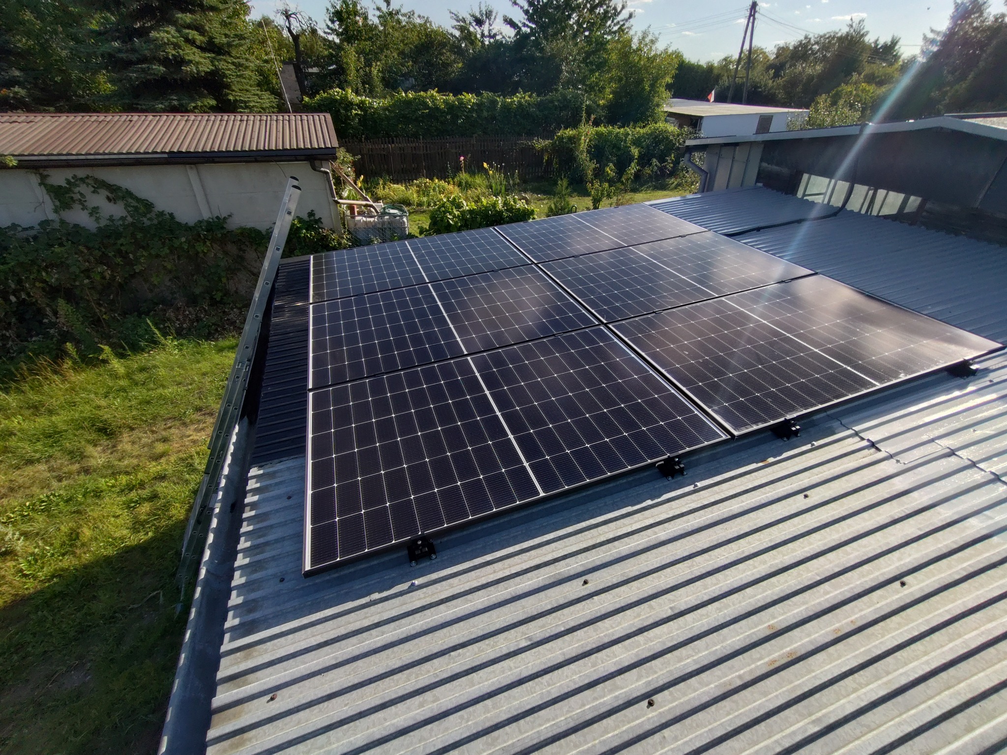 Cztery panele fotowoltaiczne zamontowane na blaszanym dachu budynku, widoczne z góry, z drabiną opartą o krawędź dachu i zielonym otoczeniem w tle.