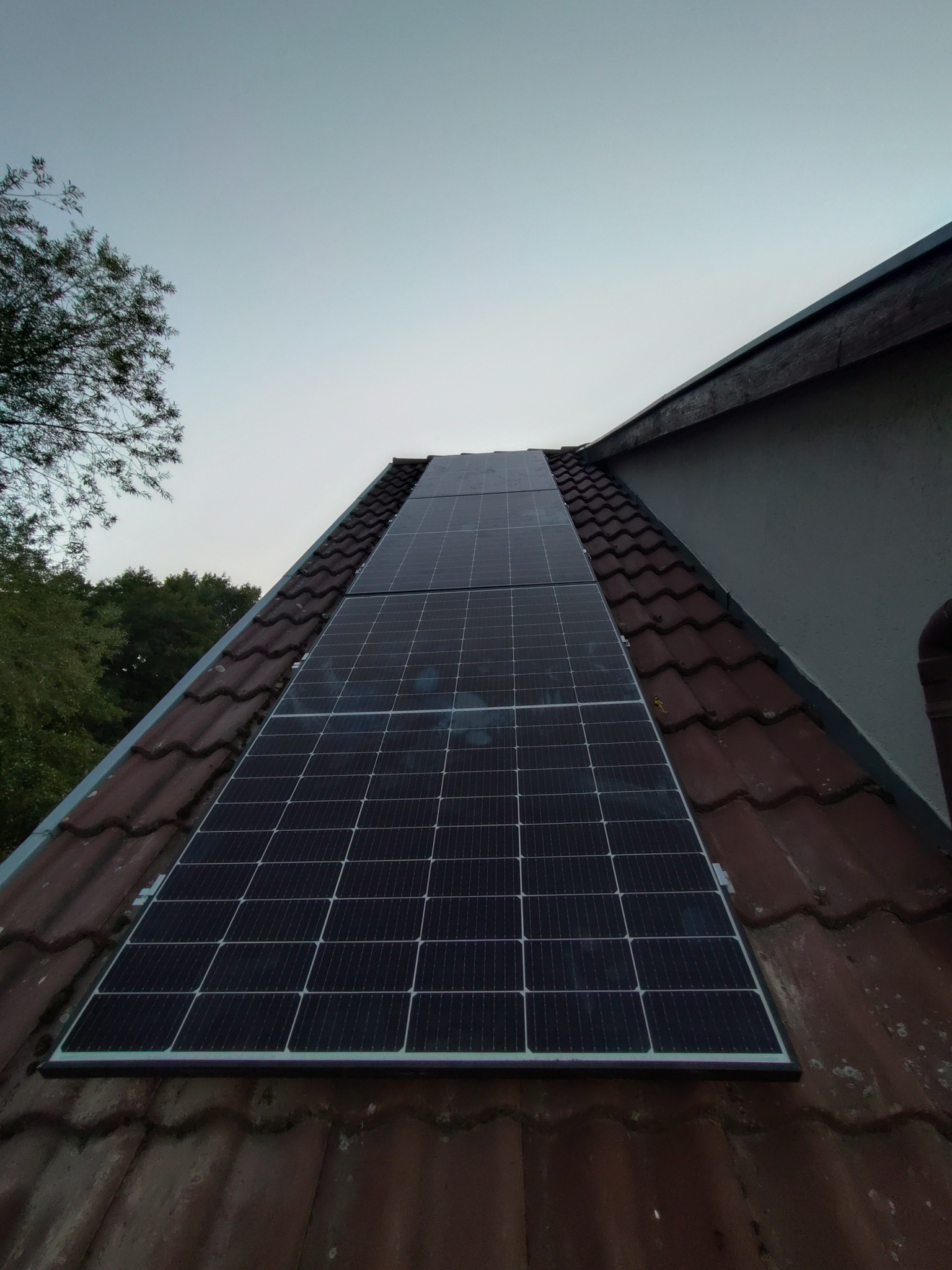 Panele fotowoltaiczne na dachu z czerwonej dachówki, widok z dołu pod kątem. Częściowo pochmurne niebo w tle. Widoczne drzewa po lewej stronie.