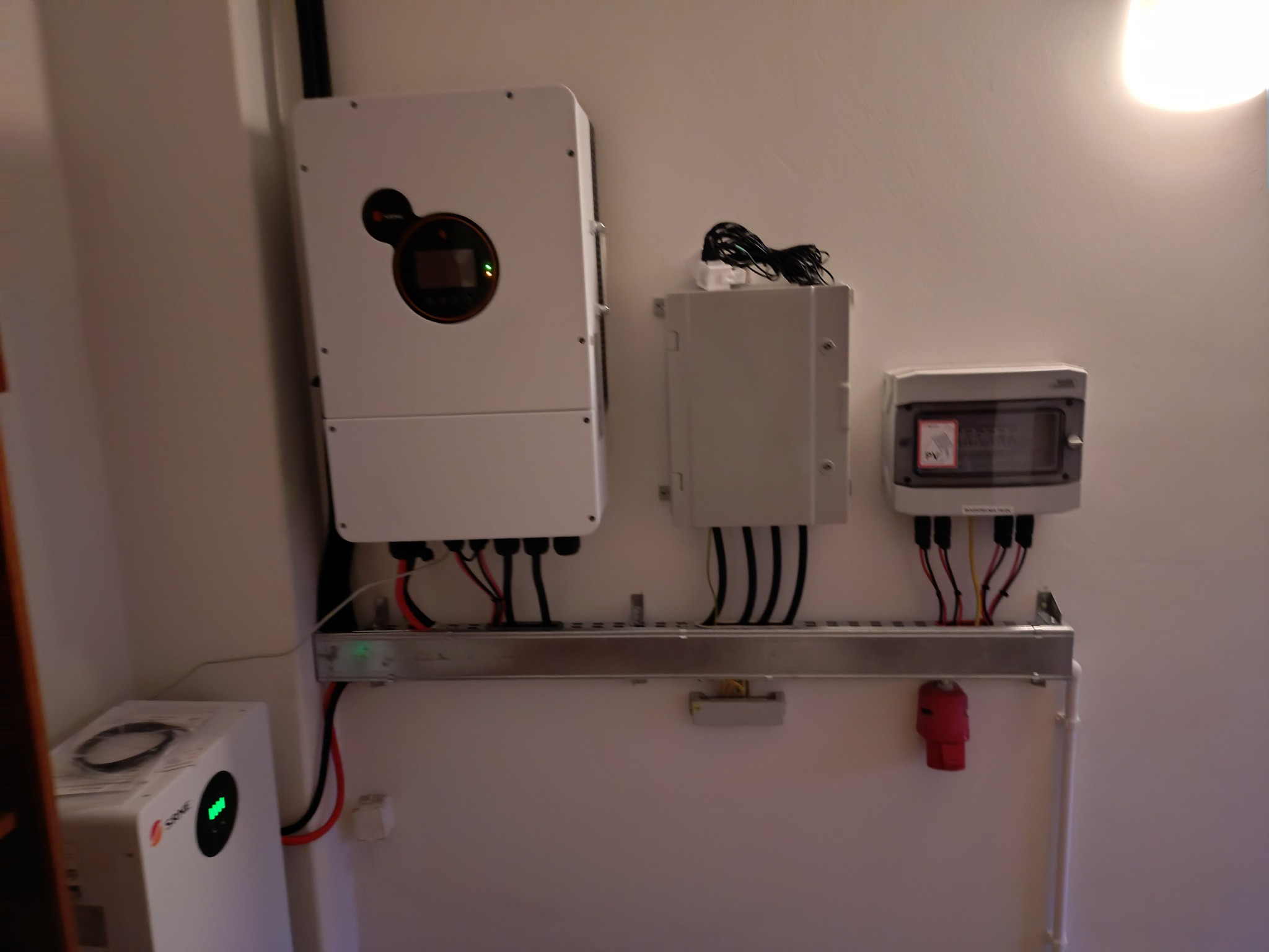 Białe urządzenia elektryczne, w tym falownik z wyświetlaczem, zamontowane na ścianie z widocznymi kablami i przewodami. Element instalacji fotowoltaicznej wewnątrz budynku.