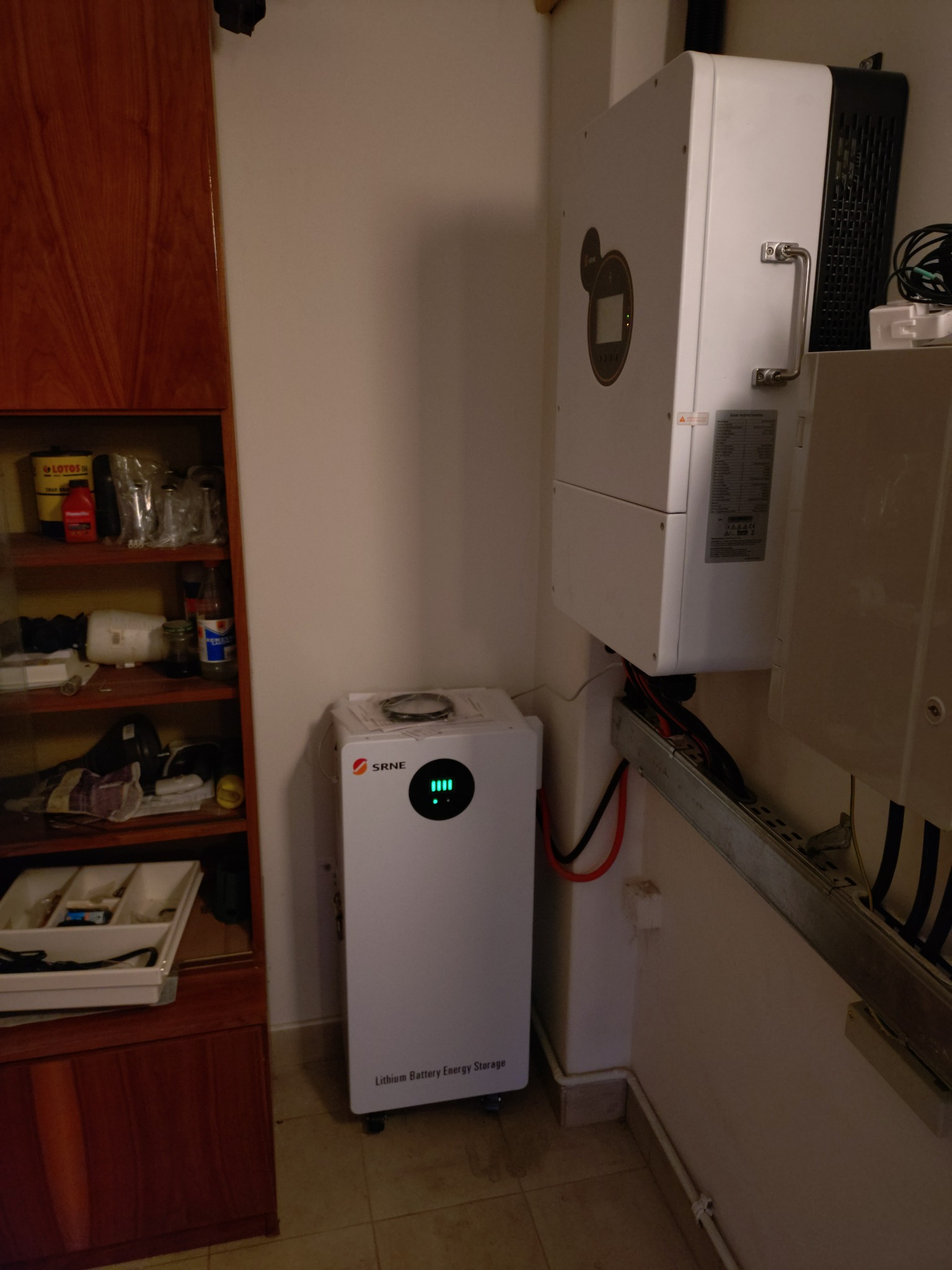 Biały magazyn energii SRNE i inwerter na ścianie w pomieszczeniu. Widoczne kable i elementy instalacji. Obok szafka z wyposażeniem.